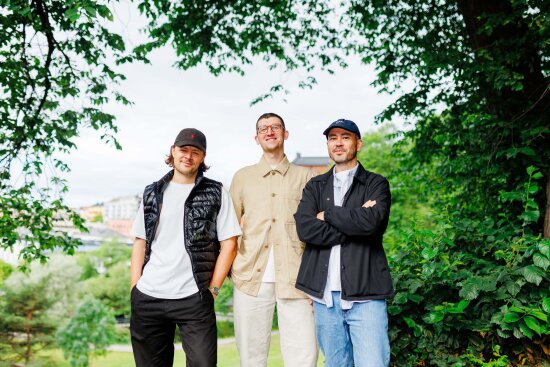 Tre menn ser smilende mot kamera, omringet av trært. Foto.