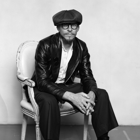 Svart- hvitt-portrett av en mann kledd i svart skinnjakke, svarte bukser og hvit skjorte sittende på en antikk stol. På hodet har han en stor sixpence.