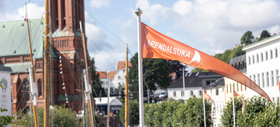 En oransje vimpel med teksten Arendalsuka med seilbåtmaster og et kirketårn i bakgrunnen.