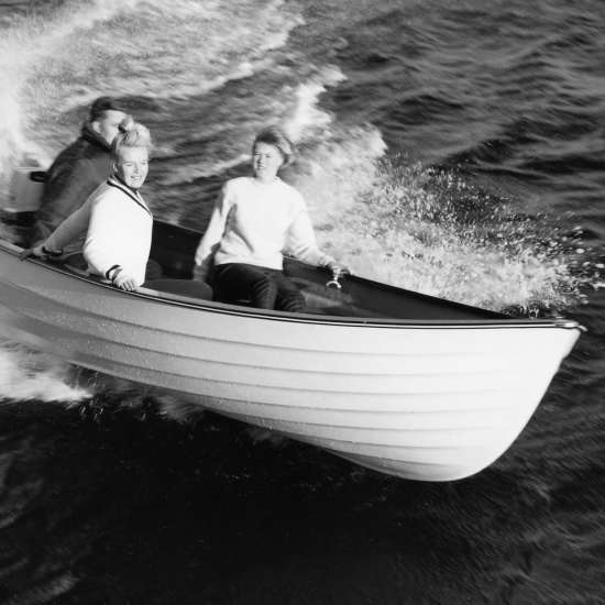 Svart-hvitt-bilde av to kvinner og en mann i en jolle med påhengsmotor.
