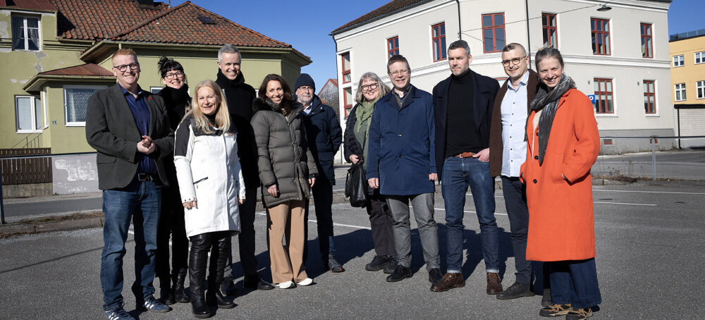 11 personer poserer på en parkeringsplass foran noen eldre bygninger. De smiler og himmelen er blå bak dem.  