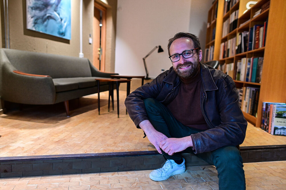 En blid mann med skjegg og briller sitter på et gulv med flere nivåer. I bakgrunnen er det en bokhylle, en sofa og en lampe. Sofaen og lampen er i 50-tallsstil. Mannens navn er Jan Erik Rossow.