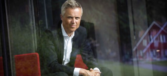 En mann med grått hår og direkte blikk, sort dressjakke og hvit skjorte, sitter i en rød stol.