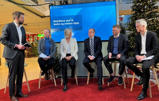 Et panel med fem deltakere som sitter på høye krakker. På venstre side står en møteleder i sort dress og hvit skjorte. På en skjerm bak panelet står teksten bedriftene skal få enklere og bedre hjelp.