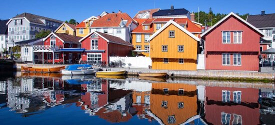 Røde og gule trehus på en bryggekai speiler seg i vannet. Ved kaia ligger det fire småbåter.