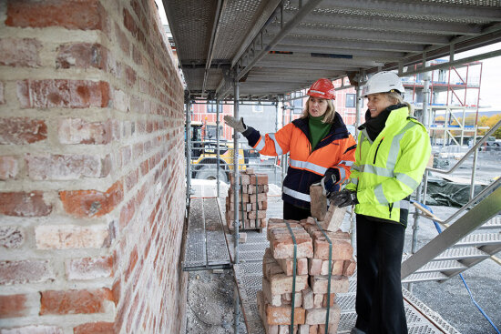 To damer ikledd verneutstyr står foran mursteinsvegg på anleggsområde.