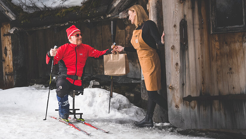 En mann i solbriller og langrennstøy mottar en take away-pose med mat av en kvinne i forkle. Mannen har et amputert bein, og sitter på en skikjelke. 