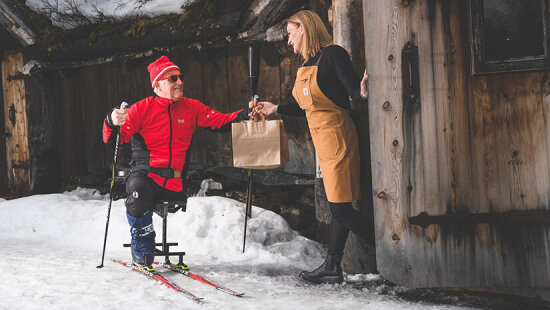 En mann i solbriller og langrennstøy mottar en take away-pose med mat av en kvinne i forkle. Mannen har et amputert bein, og sitter på en skikjelke.