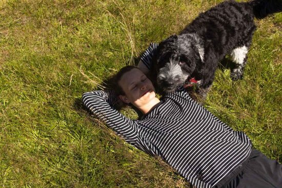 Stine ligger i gresset med amene over hodet. Hunden Tussi står over henne og ser opp i kamera.