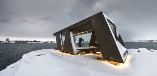 Fugleskjul med en fotograf inni som tar bilde av fugler. Tettsted i bakgrunnen. Snø på bakken.