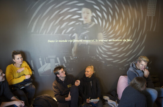 Ungdommer som sitter langs en vegg innendørs, hvor et bilde av en DJ med teksten «Dans le monde réellement renversé, le vrai est un moment du faux.» projiseres.