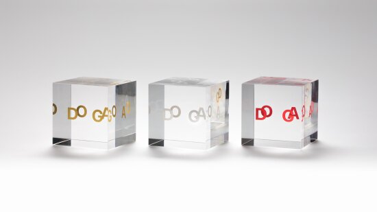 Tre DOGA-merket-plaketter; glasskuber med bokstavene D, O, G og A inni, i gull, hvitt og rødt.
