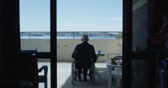 Eldre mann i rullestol sitter på en balkong og ser utover en fjord og landskap på andre siden av fjorden. Bildet tatt bakfra