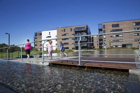 Mor med to barn som går på gangbro over vannspeil. Nybygg (lavblokker) i bakgrunnen.