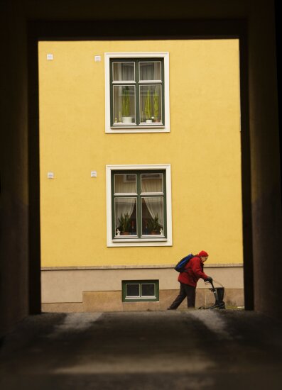 Gul bygård, i front en eldre dame i rødt med en gåstol.