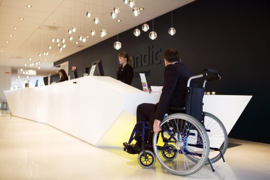 Mann i rullestol foran resepsjon på hotell. Scandic Hotel Gardermoen var hovedvinner av Innovasjonsprisen for universell utforming i 2011.