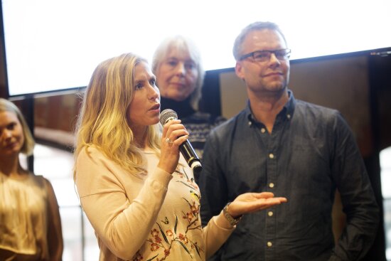 En dame står og snakker i mikrofonen. I bakgrunnen står tre andre - to damer og en mann.  Foto.