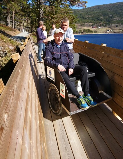 I forgrunnen en mann i rullestol. Bak stolen en annen mann og bak der igjen to menn og en dame. De tre siste ser ut som de snakker sammen. Alle står på en bro eller gangvei av tre. Til høyre er det sjø og til venstre og bak er det skogbunn og furutrær.
