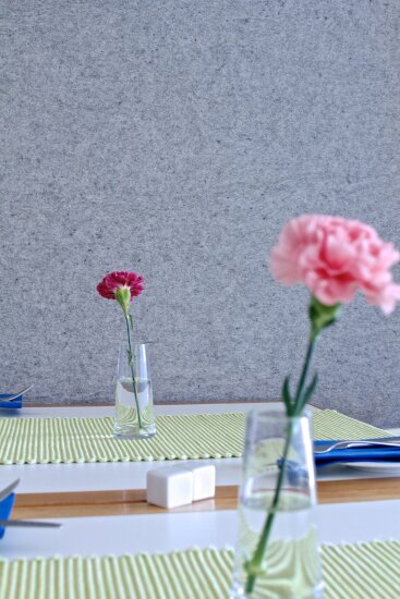 Nærbilde av vegg med overflate av feltroll. På et bord med lysegrønn løper står det to blomstervaser med en enkelt nellik i hver. Blomsten nærmest er rosa, den andre er cerisefarget.