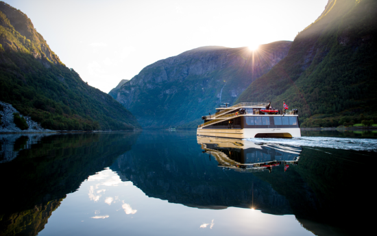Båten Vision of the Fjords på vei inn en fjord med blikkstille overflate. Høye fjell, solen ligger på fjellsiden og stråler ned over båten. Fjellene, båten, himmelen og noen skyer speiler seg i vannoverflaten.