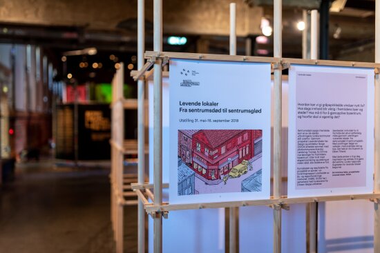 Plakater fra utstillingen Levende lokaler, satt opp i et rammeverk av tre. I bakgrunnen skimtes lys og annet innover i Hallen på DOGA.