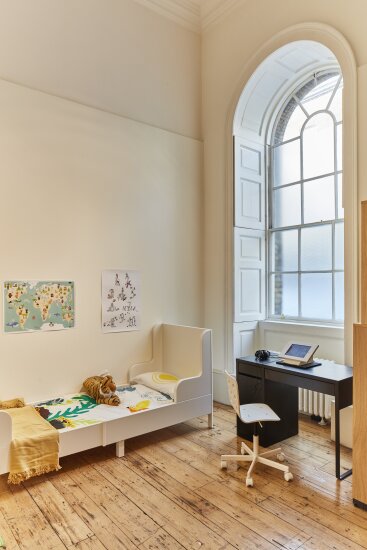 Et stort rom, høyt under taket, store, buede vinduer. En oppredd barneseng står i et hjørne og ved vinduet står det et skrivebord. På skrivebordet er det et nettbrett.