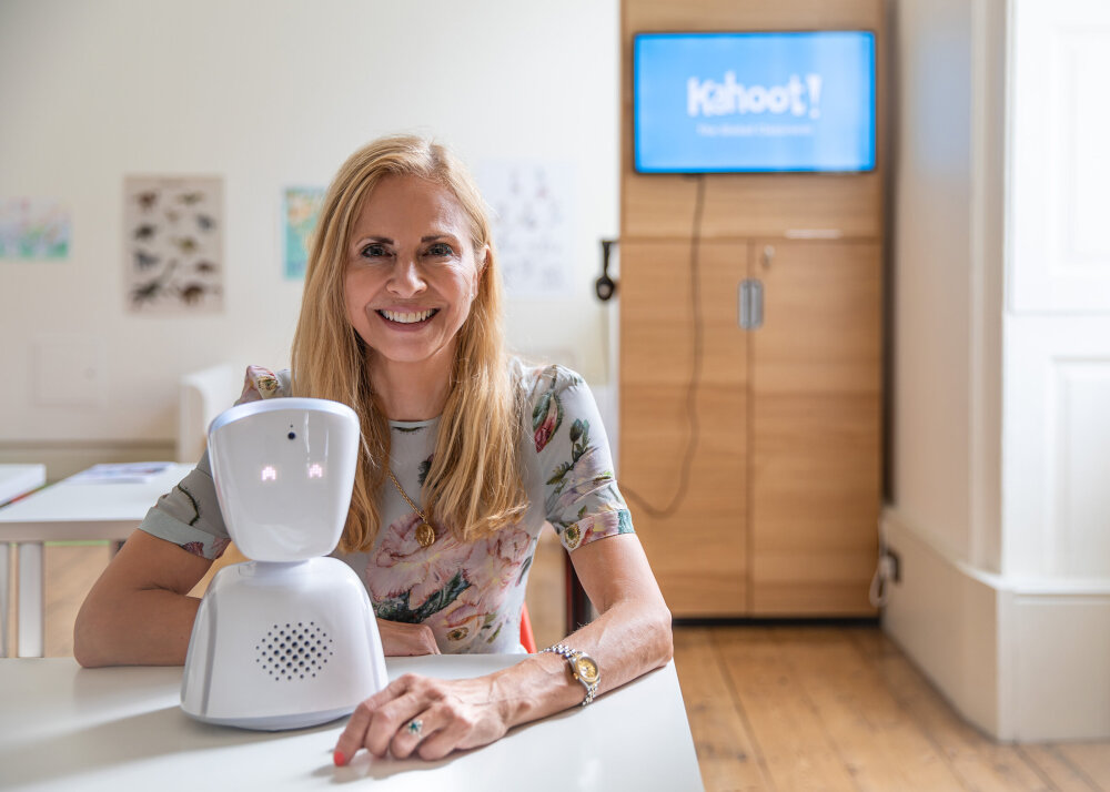 Kurator for DOGAs utstilling på London Design Biennale 2018, Onny Eikhaug. Hun holder roboten AV1 og i bakgrunnen ser vi utstillingslokalet. På veggen henger det en skjerm hvor man kan skimte at det står Kahoot.  