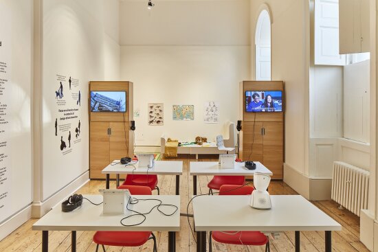 Et stort, luftig, hvitmalt rom, med høye buede vinduer. Rommet er innredet som et klasserom med fire pulter. Ved hver pult står en rød stol. På tre av pultene står det et nettbrett, og på den fjerde pulten står roboten AV1.