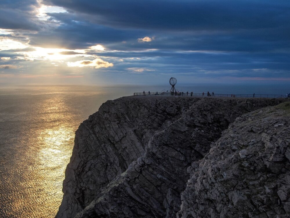 Klippe (Nordkapp-platået) med globus-installasjon ved havet i solnedgang.