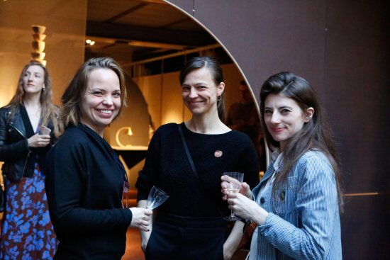 Tre damer smiler mot kameraet. To av dem holder et vinglass. I bakgrunnen til venstre står en annen dame og bak henne igjen ser vi den runde åpningen inn til et av utstillingsrommene.