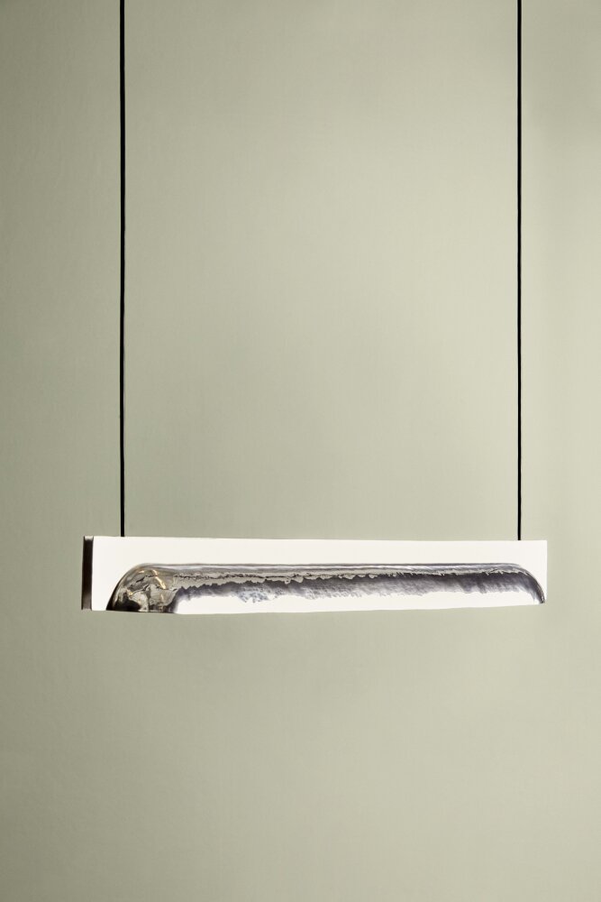En smal rektangulær lampe som henger i to lange ledninger festet i hver sin ende av rektangelet. Lampen er laget av aluminium og er design av Stine Aas. Den skal stilles ut ved den norske fellesutstillingen under Milano Design Week 2019.