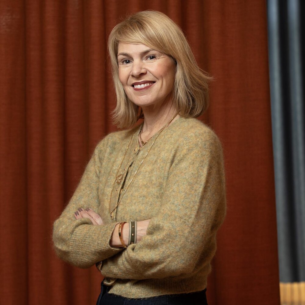 Anne smilende med blondt halvlagt hår og gulbeige jakke.