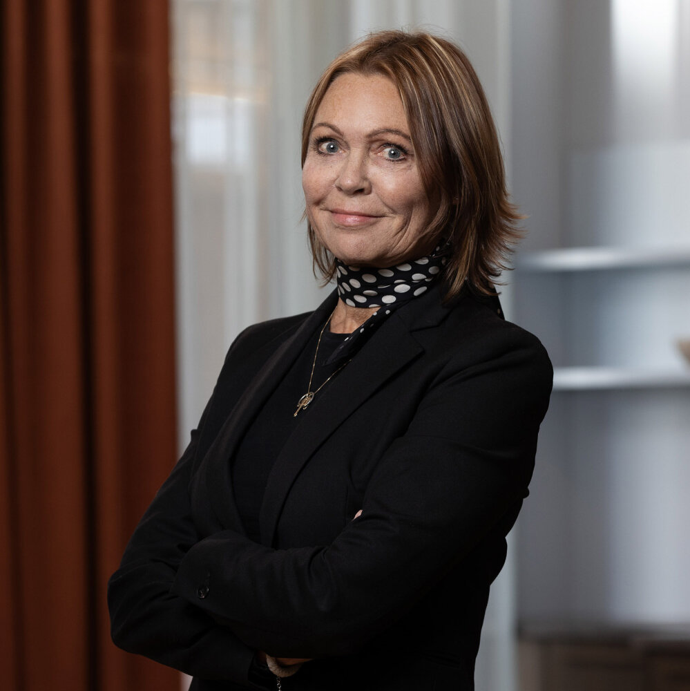 Elisabeth ser rett inn i kameraet med et direkte blikk og et lite smil. Hun har kort, mørk blondt hår, sort blazer og et lite sort skjerf med hvite prikker rundt halsen.