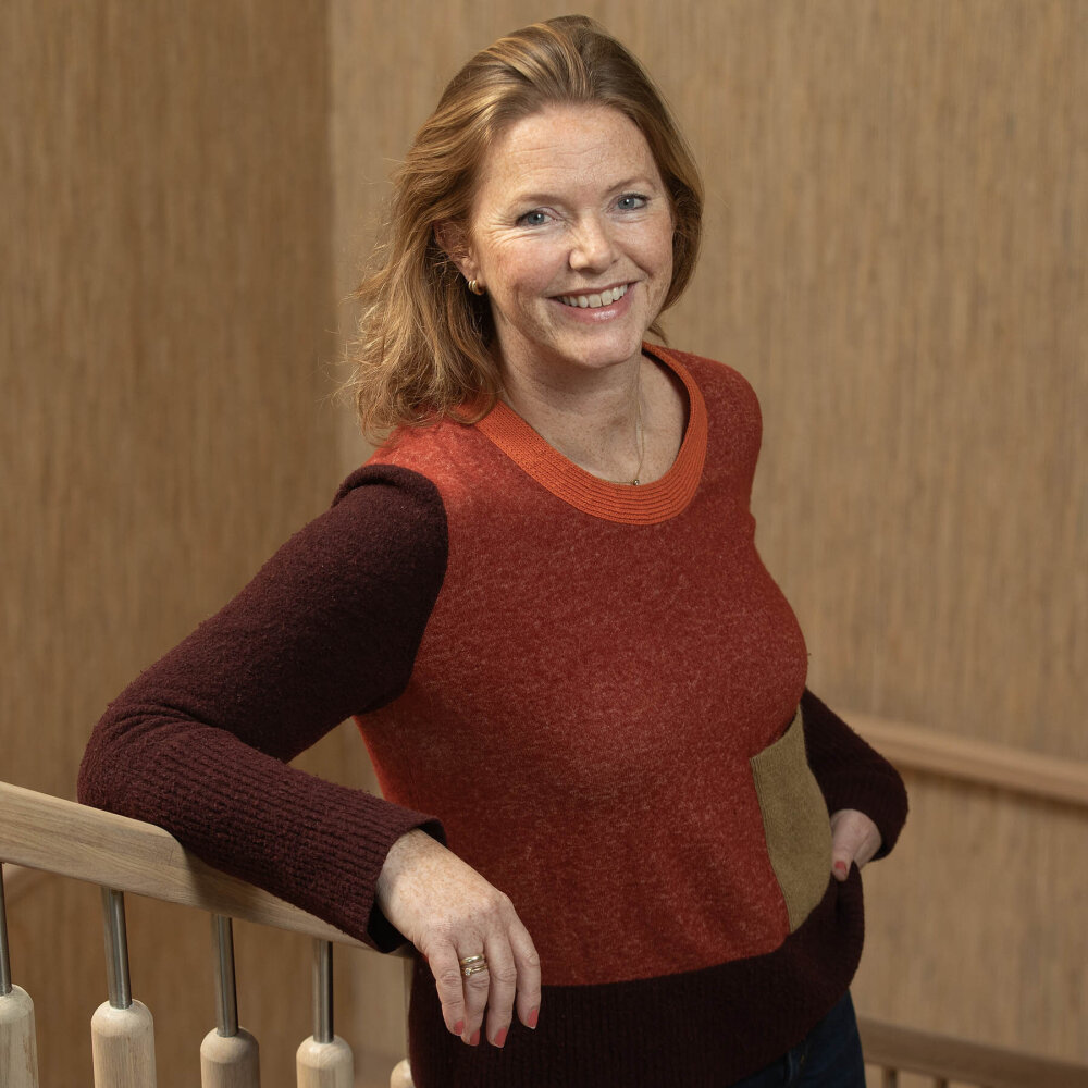 Hilde står i trappen med et stort smil. Hun har rødlig, halvlagt hår gredd bakover og en topp i ulike rødbrune og beige fargetoner. Neglene er malt i en rødoransje farge. 