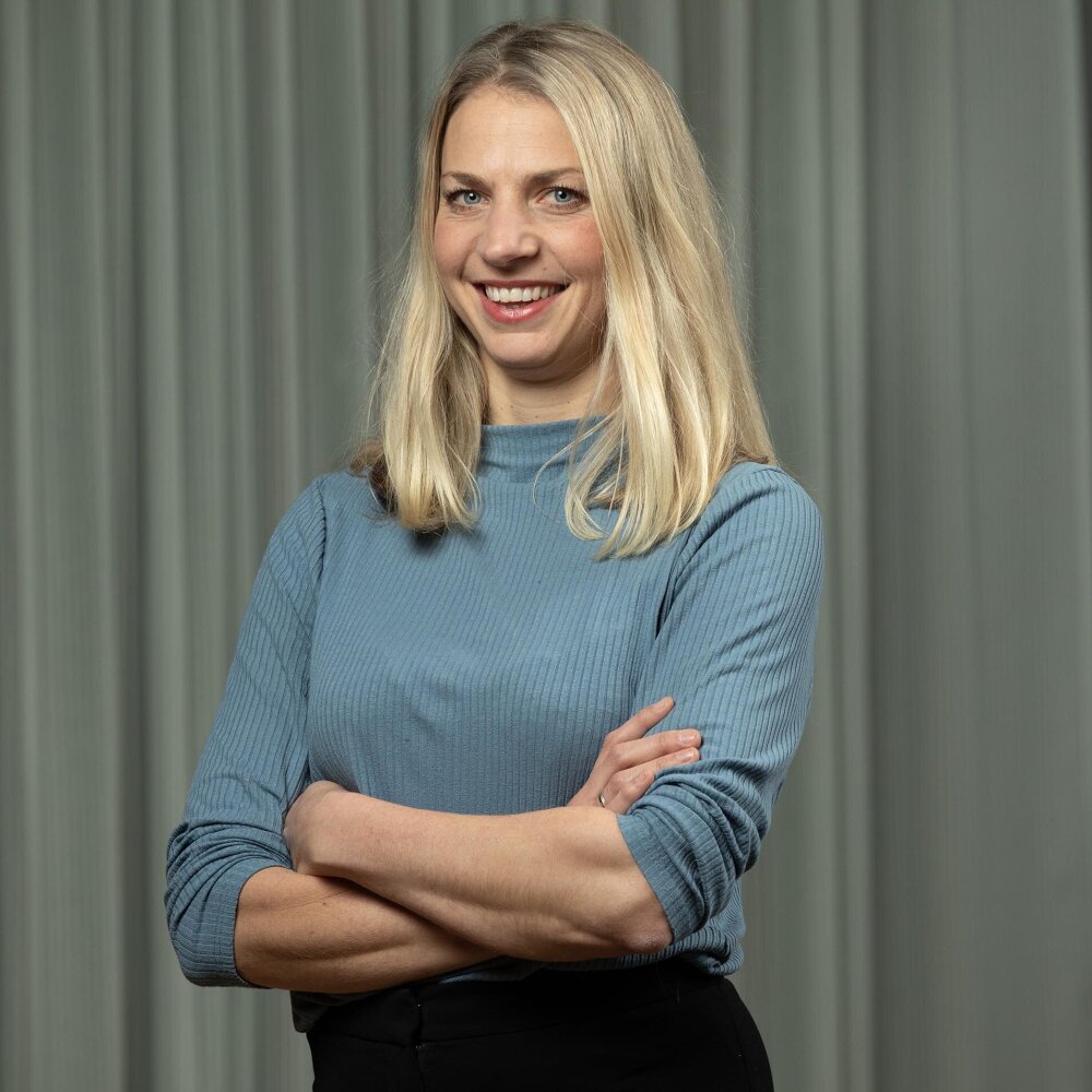 Ingeborg med armene i kors og et stort smil. Hun har halvlangt lyst hår og har på seg en dueblå genser. 