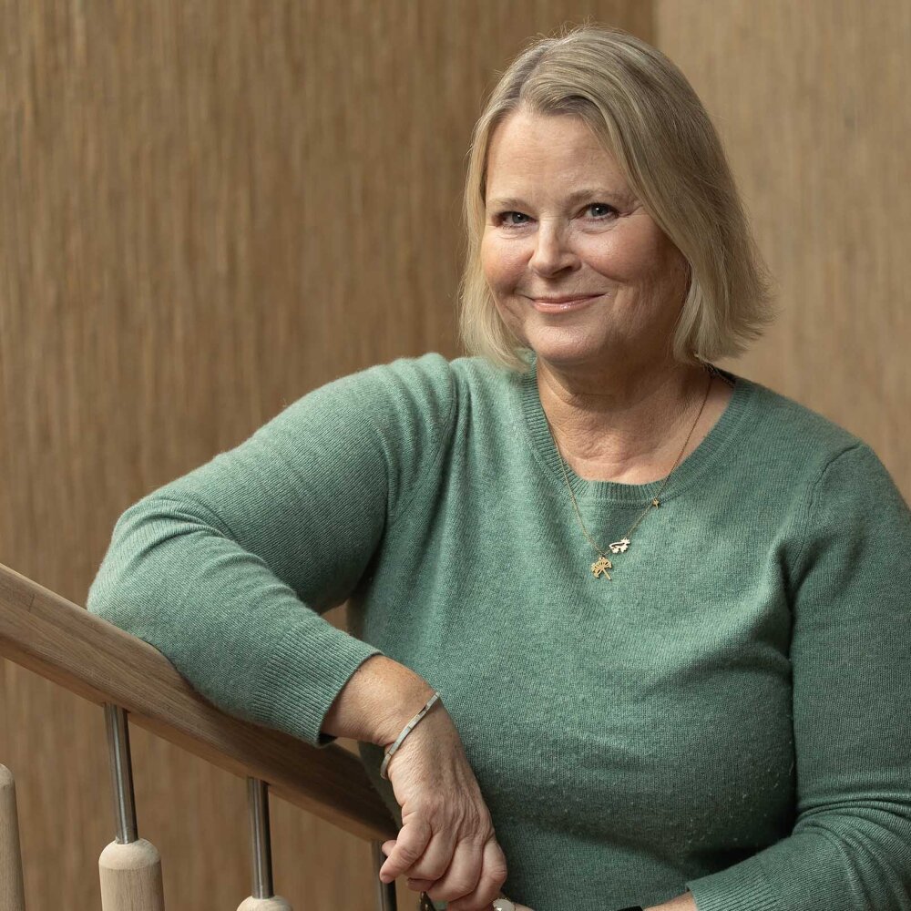 Jannicke står lent opp mot trappegelenderet med et lunt smil. Hun har på seg en blågrønn genser og et tynt gullkjede med anheng. 