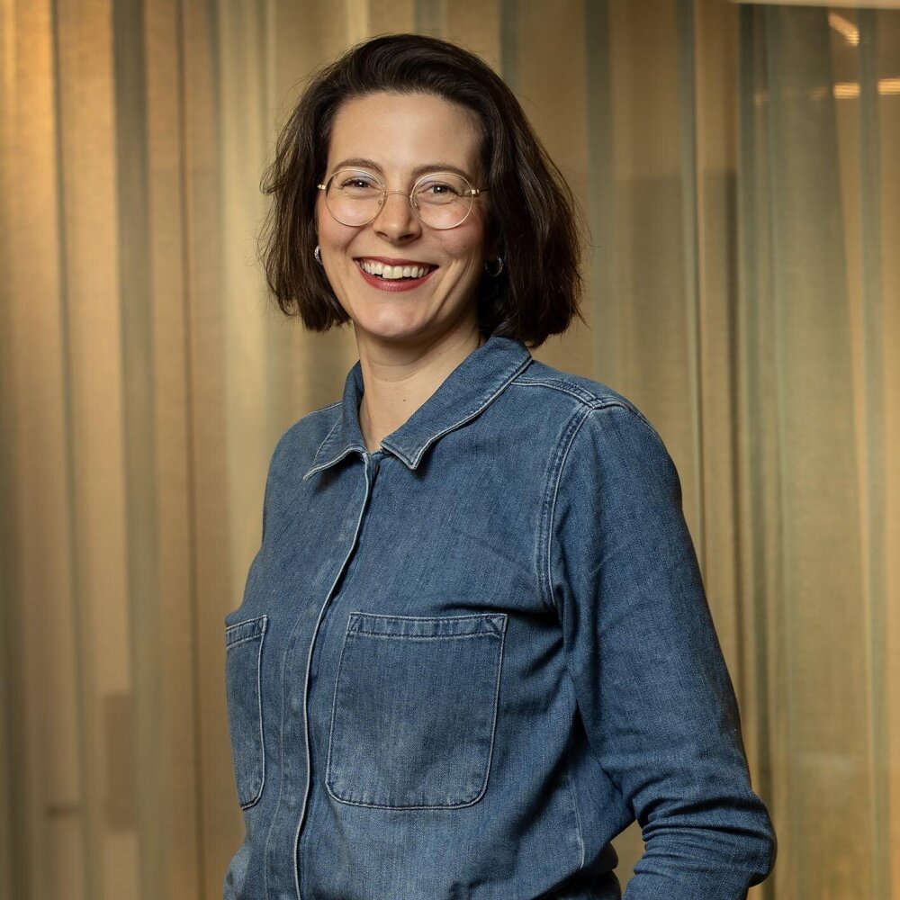 Kjersti med et varmt smil og kort, mørkt hår. Hun har biller med tynn, gylden innfatning og har på seg en blå jumpsuit i blå denim. 