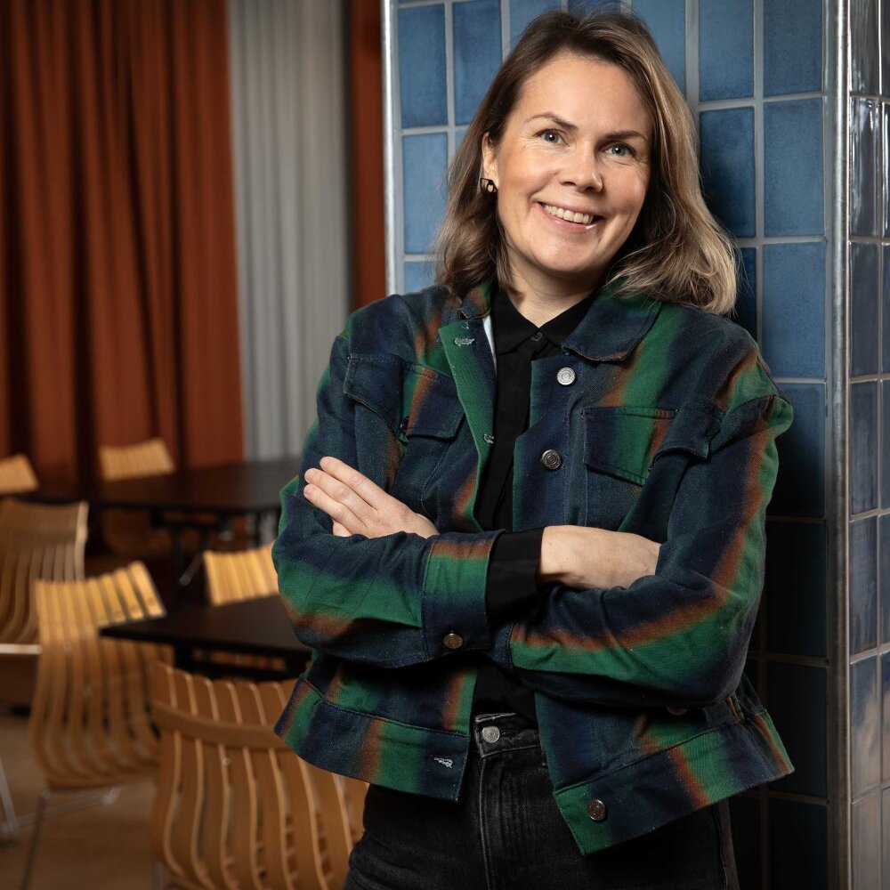 Malin har halvlangt mørkt hår, grønnrutete jakke og holder armene i kors. Hun smiler mot kamera.