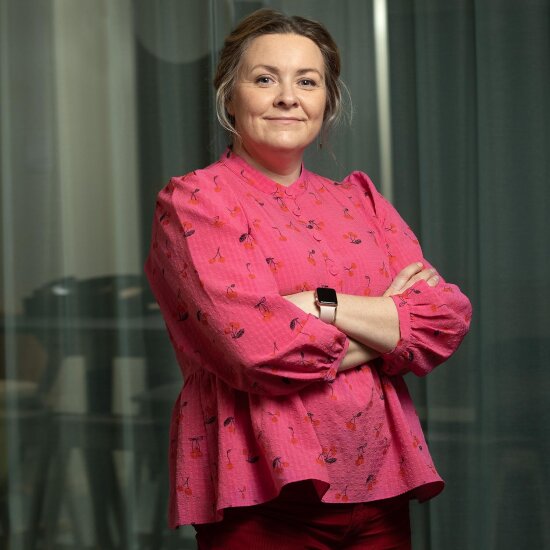 Marianne ser vennlig inn i kameraet. Hun har oppsatt, blondt hår, med litt løst hår hengende ned på hver side av ansiktet. Hun har på seg en rosa bluse, og på armen har hun en smartklokke med pudderrosa rem.