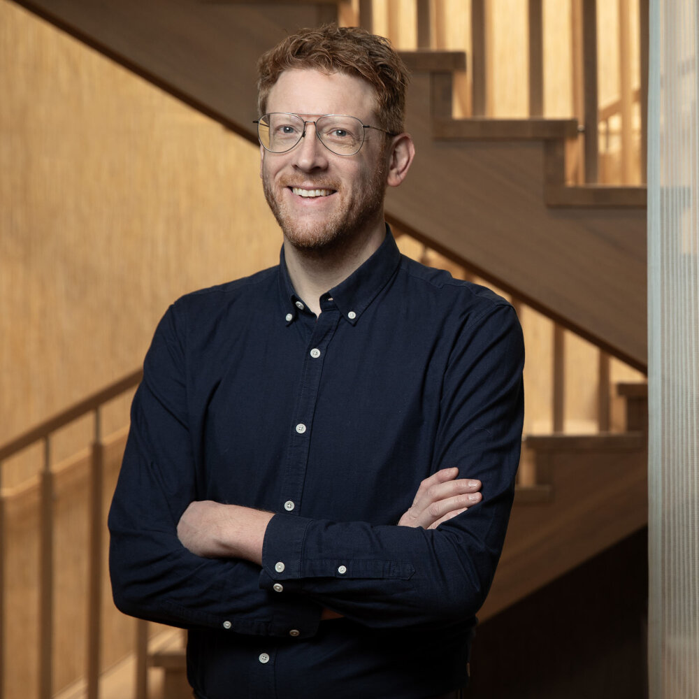Med kort, rødt hår og briller står Martin med et vennlig smil, Han har på seg en marineblå skjorte og står med armene i kors foran en trapp.  