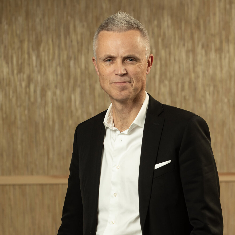 Tor Inge med et lite smil, kort, grått hår, sort dressjakke og hvit, åpen skjorte. 