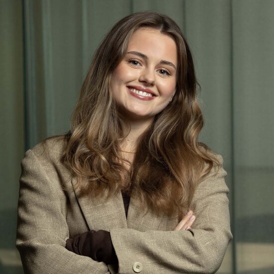 Una med et stort smil og hodet litt på skakke. Hun har langt, blondt hår og beige dressjakke.