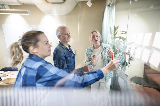 Tre kollegaer står vendt mot en glasstavle i livlig diskusjon.