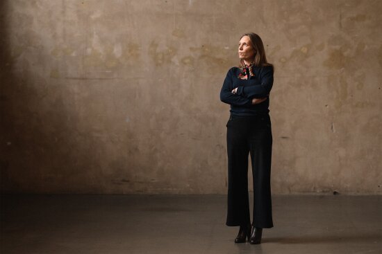 Administrerende direktør i DOGA, Trude Gomnæs Ugelstad, ser ut mot lyset i hallen på DOGA. Hun har på seg blå bukse og bluse, og et silkeskjerf i halsen. Hun har armene i kors.