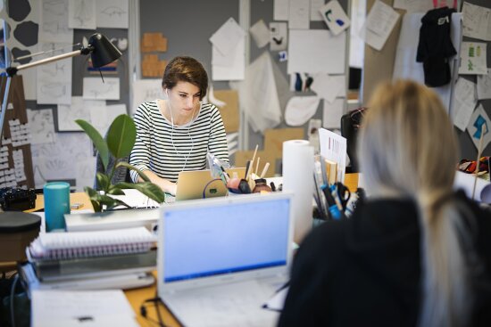 Jente med hodetelefoner som arbeider på bærbar datamaskin i et kontor-/verksted-miljø.
