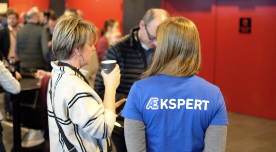 To kvinner snakker sammen med ryggen til. Den ene har t-skjørte med påskriften «ÆKSPERT».