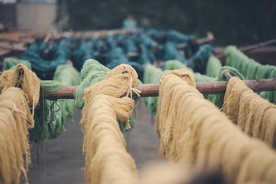 Fargede tråder i sennepsgul, grønn og sjøgrønn henger til tørk på trestammer. Foto