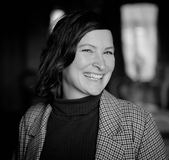 Katja smiler blidt mot kamera, med rutete jakke og mørkt hår. Foto.