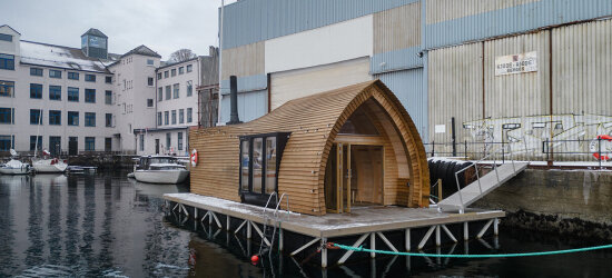 En badstuebygning med buet tak ligger fortøyd til en brygge ved en lagerbygning. Et tykt grønt fortøyningstau går fra badstuen til land.