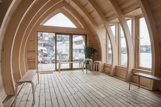 Et rom med høyreiste, bueformede limtrebjelker. Endeveggen er vinduer og en skyvedør i glass.